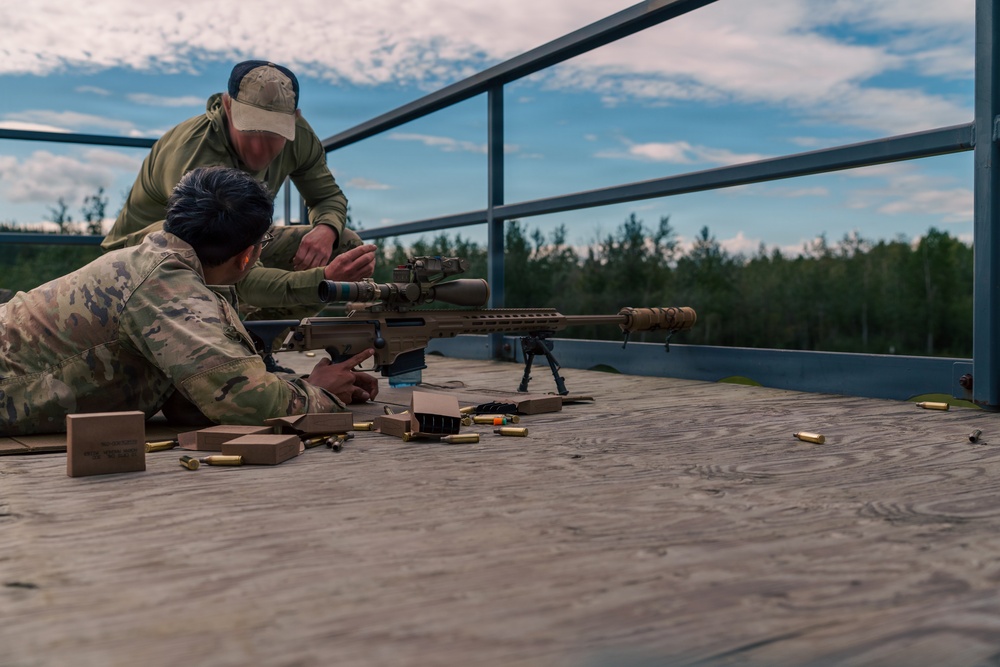 Two individuals in military/tactical attire are on an elevated wooden platform at what appears to be a shooting range. One person is in a prone position operating a large precision rifle with a bipod and scope, while a second person stands behind providing instruction or guidance. Spent brass casings and ammunition boxes are visible on the platform surface.
