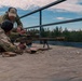 Two individuals in military/tactical attire are on an elevated wooden platform at what appears to be a shooting range. One person is in a prone position operating a large precision rifle with a bipod and scope, while a second person stands behind providing instruction or guidance. Spent brass casings and ammunition boxes are visible on the platform surface.