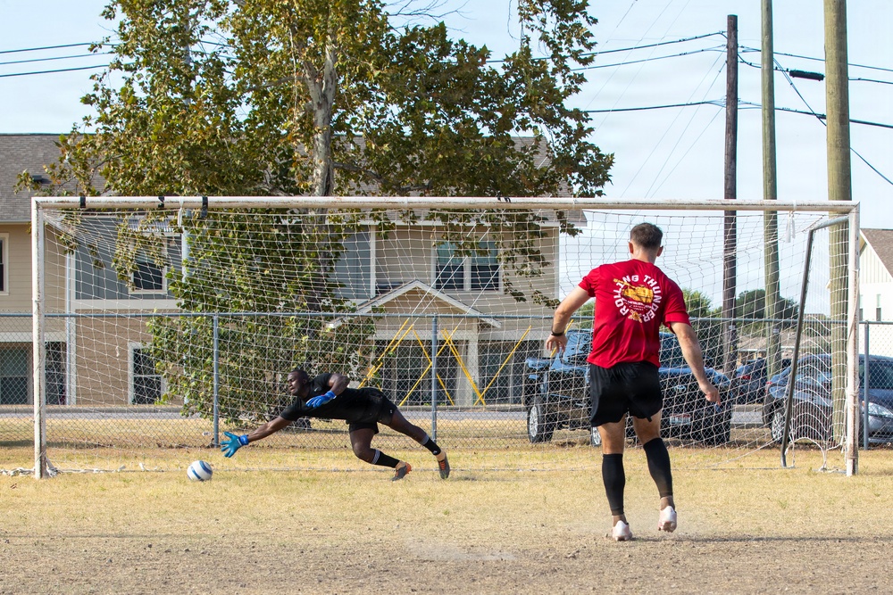 Two players are engaged in a soccer penalty kick scenario on a dry grass field. A goalkeeper in black is diving to his left attempting to save a shot, while a player in a red shirt labeled 'Rolling Thunder' stands near the goal having just kicked the ball. The setting appears to be a recreational or community field with residential buildings and parked cars visible in the background.