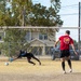 Two players are engaged in a soccer penalty kick scenario on a dry grass field. A goalkeeper in black is diving to his left attempting to save a shot, while a player in a red shirt labeled 'Rolling Thunder' stands near the goal having just kicked the ball. The setting appears to be a recreational or community field with residential buildings and parked cars visible in the background.