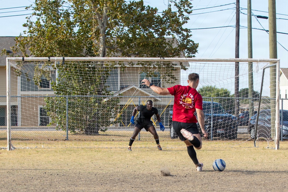Two individuals are practicing soccer on a dry, dirt field with a full-size goal. A player in a red shirt is in the process of kicking a blue and white soccer ball toward the goal, while a goalkeeper dressed in all black with blue gloves is positioned in front of the net in a ready stance. The setting appears to be a casual community or recreational field surrounded by residential buildings and trees.