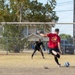 Two individuals are practicing soccer on a dry, dirt field with a full-size goal. A player in a red shirt is in the process of kicking a blue and white soccer ball toward the goal, while a goalkeeper dressed in all black with blue gloves is positioned in front of the net in a ready stance. The setting appears to be a casual community or recreational field surrounded by residential buildings and trees.