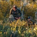 Two individuals in tactical/military gear are positioned within dense autumn foliage, partially concealed by shrubs and brush. The standing individual appears to be operating a handheld device while wearing a plate carrier vest, while the second individual is lower in the vegetation wearing a cap and headset. The setting suggests a field operation, training exercise, or reconnaissance activity in a natural outdoor environment.