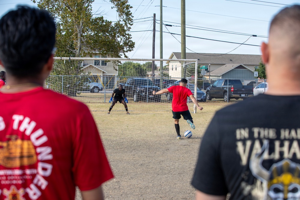 A casual outdoor soccer match is taking place on a dry, dirt field. A player in a red shirt is about to take a shot on goal while a goalkeeper in black with blue gloves prepares to defend. Two spectators in the foreground — one wearing a red 'Thunder' shirt and one wearing a black 'Valhalla' shirt — watch the action from behind.