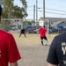 A casual outdoor soccer match is taking place on a dry, dirt field. A player in a red shirt is about to take a shot on goal while a goalkeeper in black with blue gloves prepares to defend. Two spectators in the foreground — one wearing a red 'Thunder' shirt and one wearing a black 'Valhalla' shirt — watch the action from behind.