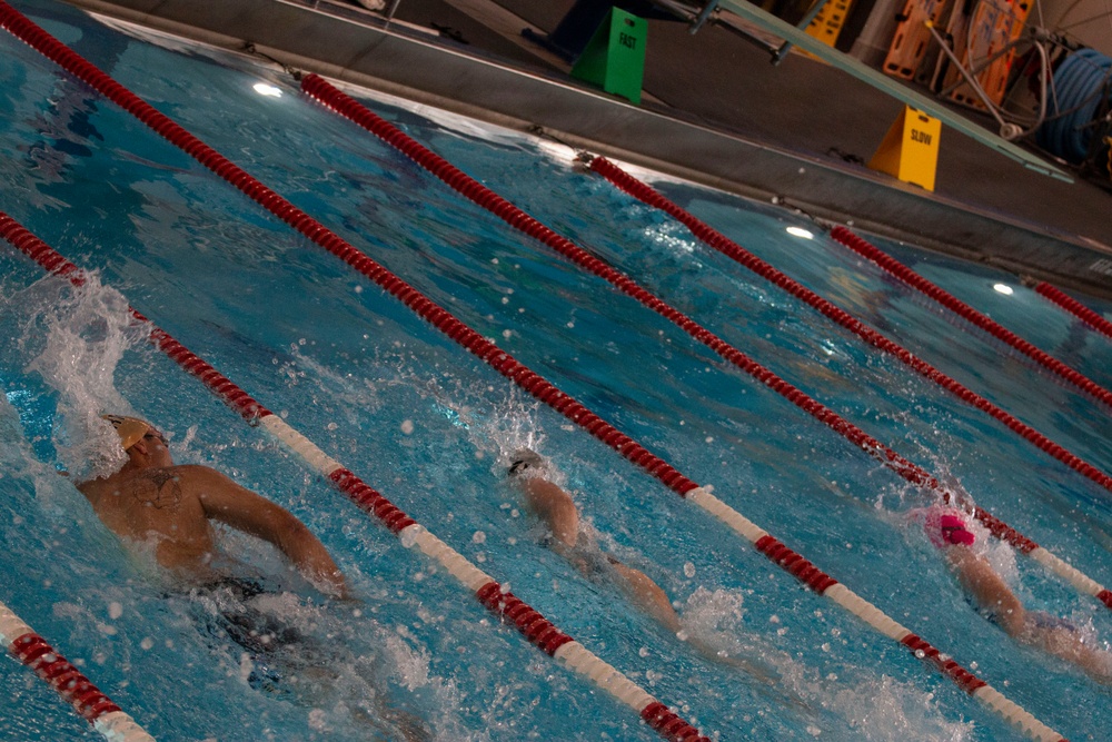 An indoor swimming pool with multiple lanes separated by red and white lane dividers. Three swimmers are actively swimming in adjacent lanes, moving away from the camera. Poolside signage indicates lane speed designations, with a green 'FAST' sign and a yellow 'SLOW' sign visible at the far end.