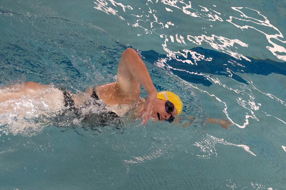 A swimmer is captured mid-stroke performing the freestyle (front crawl) technique in an indoor pool. The swimmer is wearing a yellow swim cap and purple-tinted goggles, with their head turned to the side for a breath. The image shows significant water turbulence and splash around the swimmer, indicating active, vigorous movement.