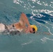 A swimmer is captured mid-stroke performing the freestyle (front crawl) technique in an indoor pool. The swimmer is wearing a yellow swim cap and purple-tinted goggles, with their head turned to the side for a breath. The image shows significant water turbulence and splash around the swimmer, indicating active, vigorous movement.