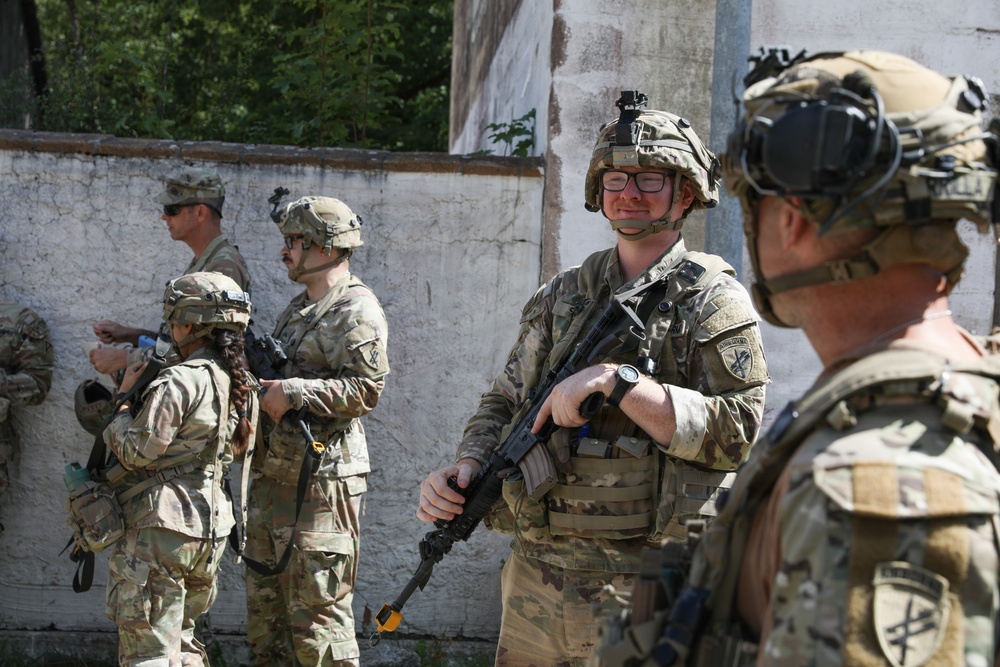 A group of military personnel in OCP (Operational Camouflage Pattern) uniforms are gathered near a weathered concrete wall in what appears to be a training exercise or operational briefing. The soldiers are equipped with helmets, body armor, and rifles, with at least one soldier wearing glasses and a helmet-mounted camera. The setting suggests an urban training environment or a forward operating area.
