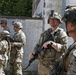 A group of military personnel in OCP (Operational Camouflage Pattern) uniforms are gathered near a weathered concrete wall in what appears to be a training exercise or operational briefing. The soldiers are equipped with helmets, body armor, and rifles, with at least one soldier wearing glasses and a helmet-mounted camera. The setting suggests an urban training environment or a forward operating area.