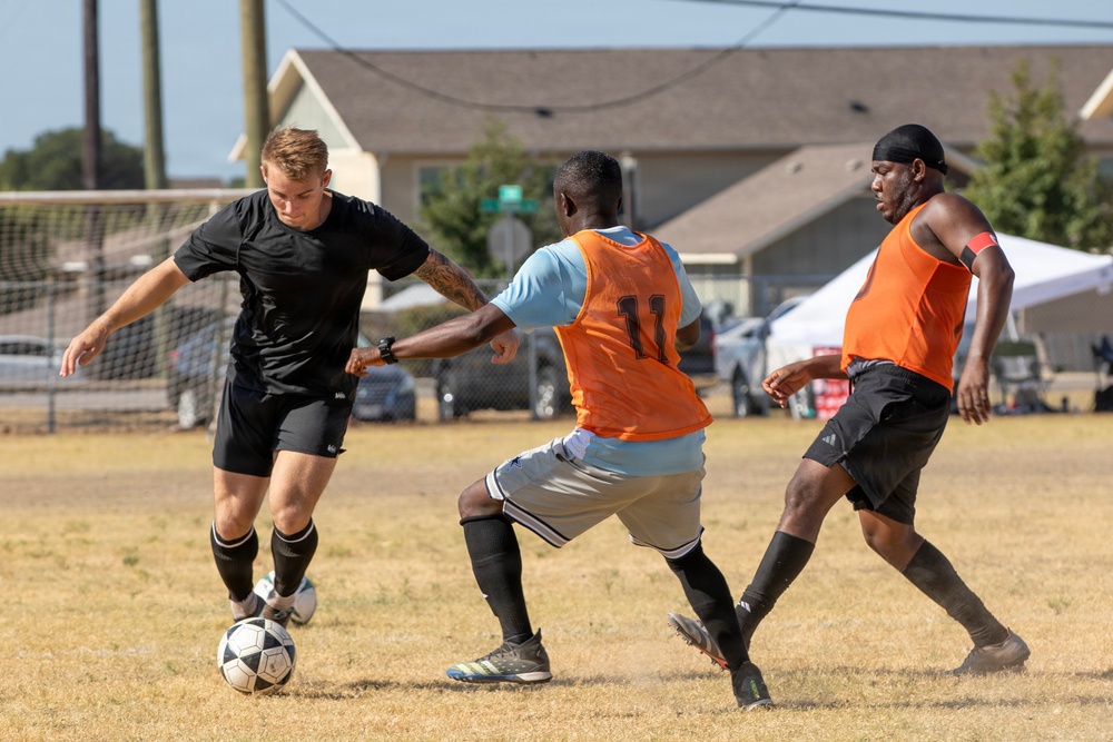 Three male soccer players are engaged in active gameplay on a dry grass field. A player in black is dribbling the ball while being defended by two players wearing orange bibs over light blue shirts. The match appears to be a recreational or amateur outdoor game, with residential buildings and parked vehicles visible in the background.