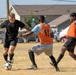 Three male soccer players are engaged in active gameplay on a dry grass field. A player in black is dribbling the ball while being defended by two players wearing orange bibs over light blue shirts. The match appears to be a recreational or amateur outdoor game, with residential buildings and parked vehicles visible in the background.