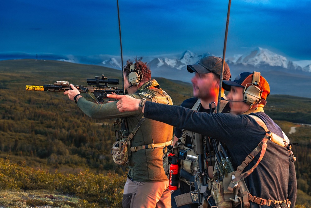 Three individuals are positioned on an elevated outdoor terrain with a mountainous, snow-capped backdrop, likely in Alaska or a similar northern region. One person is actively aiming a suppressed rifle equipped with a scope, while the other two observe. All individuals are wearing tactical hearing protection and carrying gear-laden vests or packs.