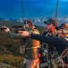 Three individuals are positioned on an elevated outdoor terrain with a mountainous, snow-capped backdrop, likely in Alaska or a similar northern region. One person is actively aiming a suppressed rifle equipped with a scope, while the other two observe. All individuals are wearing tactical hearing protection and carrying gear-laden vests or packs.