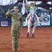 A U.S. military service member in OCP (Operational Camouflage Pattern) uniform stands at attention rendering a salute during a rodeo opening ceremony. Behind him, a rodeo queen on a white horse carries a flag while riding through the arena. The event appears to take place in Altus, Oklahoma, based on visible signage.