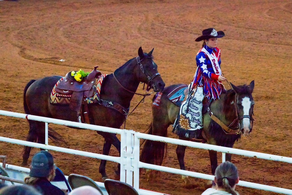 A young woman dressed in a patriotic red, white, and blue outfit with stars rides a dark horse in a rodeo arena, leading a second saddled horse beside her. The saddle blanket on her horse reads 'Great Plains Stampede Rodeo,' identifying the event. Spectators are visible in the foreground watching from behind a white fence rail.