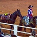 A young woman dressed in a patriotic red, white, and blue outfit with stars rides a dark horse in a rodeo arena, leading a second saddled horse beside her. The saddle blanket on her horse reads 'Great Plains Stampede Rodeo,' identifying the event. Spectators are visible in the foreground watching from behind a white fence rail.