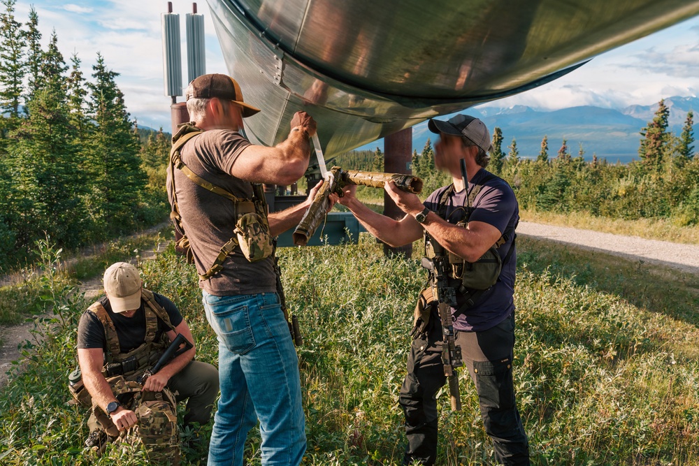 Three armed individuals in tactical gear are gathered beneath a large elevated pipeline in a forested, mountainous setting. Two standing individuals appear to be handling or attaching something to the underside of the pipeline, while a third crouches nearby. All individuals have their faces obscured/blurred in the image.