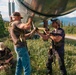 Three armed individuals in tactical gear are gathered beneath a large elevated pipeline in a forested, mountainous setting. Two standing individuals appear to be handling or attaching something to the underside of the pipeline, while a third crouches nearby. All individuals have their faces obscured/blurred in the image.