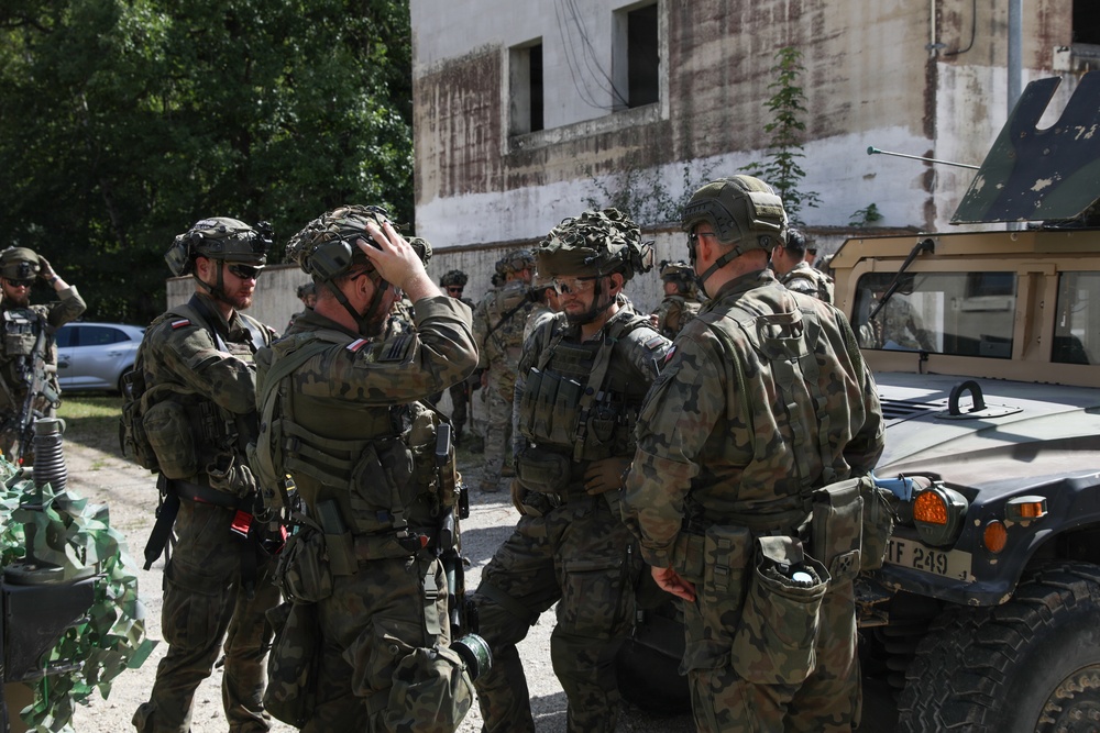 A group of soldiers in camouflage uniforms are gathered near a military Humvee vehicle, appearing to conduct a briefing or equipment check. The soldiers are wearing full combat gear including helmets with night-vision mounts, body armor, and tactical equipment. The setting appears to be a training exercise or operational staging area near a deteriorated building.