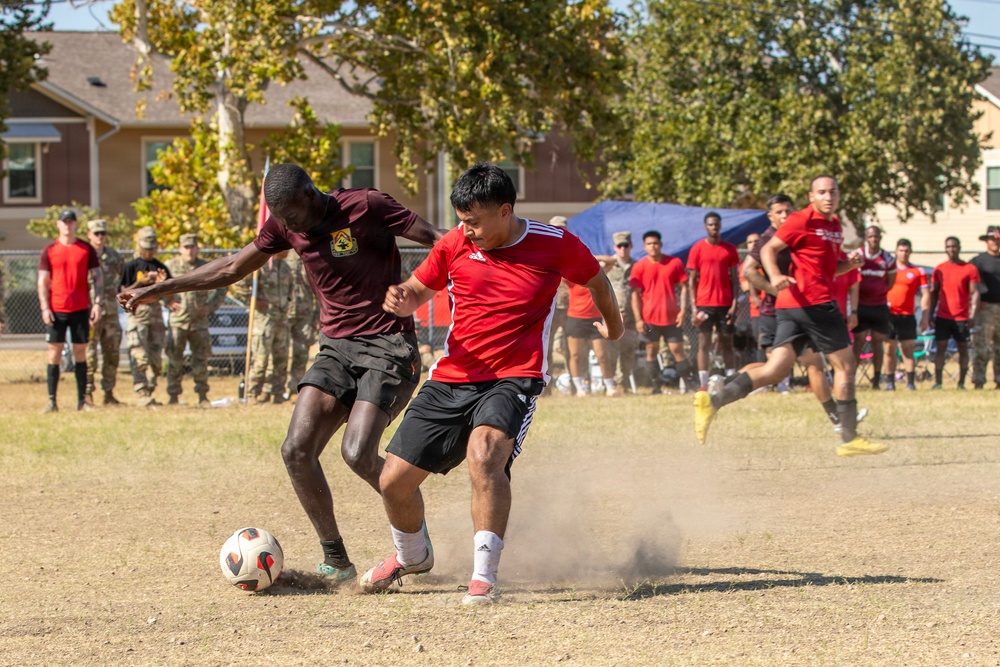 Two soccer players are competing for the ball on a dry, dusty dirt field, with one player wearing a maroon shirt and the other in a red Adidas jersey. A large group of spectators, many appearing to be in military uniforms and red athletic wear, line the sidelines watching the match. The action shot captures dust being kicked up during the intense one-on-one challenge.