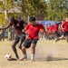 Two soccer players are competing for the ball on a dry, dusty dirt field, with one player wearing a maroon shirt and the other in a red Adidas jersey. A large group of spectators, many appearing to be in military uniforms and red athletic wear, line the sidelines watching the match. The action shot captures dust being kicked up during the intense one-on-one challenge.