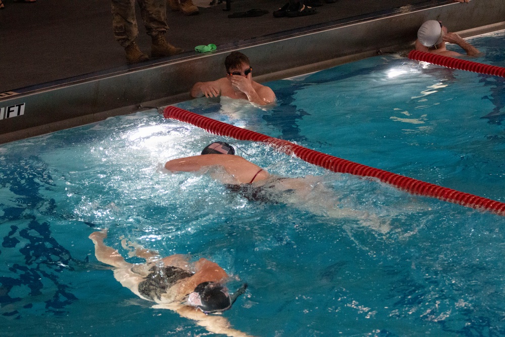 The image shows an indoor swimming pool with multiple swimmers in separate lanes divided by red lane dividers. At least three swimmers are visible actively swimming or resting at the wall, with one swimmer wearing a cap featuring what appears to be a US flag emblem. Observers in military-style boots and camouflage pants are visible standing at the pool deck in the background.