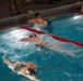 The image shows an indoor swimming pool with multiple swimmers in separate lanes divided by red lane dividers. At least three swimmers are visible actively swimming or resting at the wall, with one swimmer wearing a cap featuring what appears to be a US flag emblem. Observers in military-style boots and camouflage pants are visible standing at the pool deck in the background.
