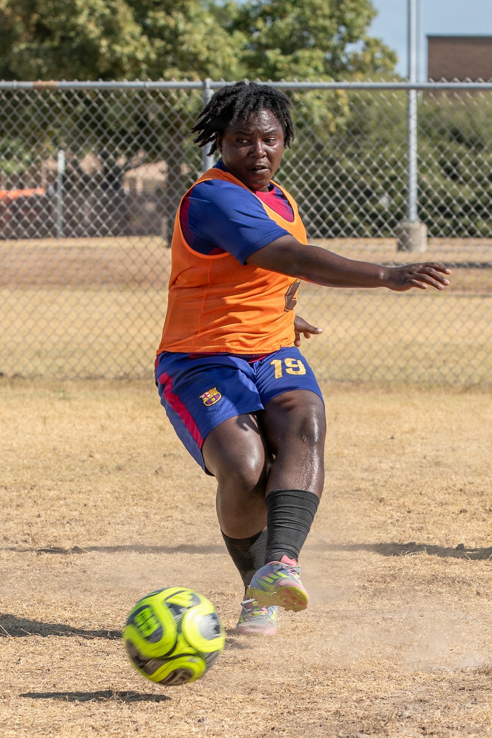 A soccer player wearing number 19 in a blue FC Barcelona kit with an orange training bib is captured mid-kick on a dry, dusty outdoor field. The player has dreadlocks, is visibly sweating, and is in a dynamic kicking stance with a yellow-green soccer ball just in front of their foot. The background shows a chain-link fence and trees, suggesting a recreational or training facility.