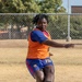 A soccer player wearing number 19 in a blue FC Barcelona kit with an orange training bib is captured mid-kick on a dry, dusty outdoor field. The player has dreadlocks, is visibly sweating, and is in a dynamic kicking stance with a yellow-green soccer ball just in front of their foot. The background shows a chain-link fence and trees, suggesting a recreational or training facility.
