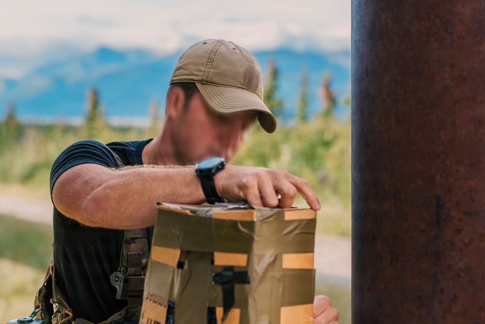 A man wearing a tan baseball cap, black t-shirt, and tactical gear is leaning over a olive-drab colored package or container near a large rusty metal pole or pipe. His face has been intentionally blurred, suggesting privacy protection. The background shows a mountainous, forested landscape under a partly cloudy sky.