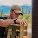 A man wearing a tan baseball cap, black t-shirt, and tactical gear is leaning over a olive-drab colored package or container near a large rusty metal pole or pipe. His face has been intentionally blurred, suggesting privacy protection. The background shows a mountainous, forested landscape under a partly cloudy sky.