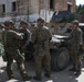 A group of soldiers in camouflage uniforms are gathered around military vehicles, including what appears to be a HMMWV (Humvee), in an urban environment near a deteriorated building. The soldiers appear to be conducting a briefing or coordination activity. Shoulder patches visible on some soldiers suggest Polish military affiliation.
