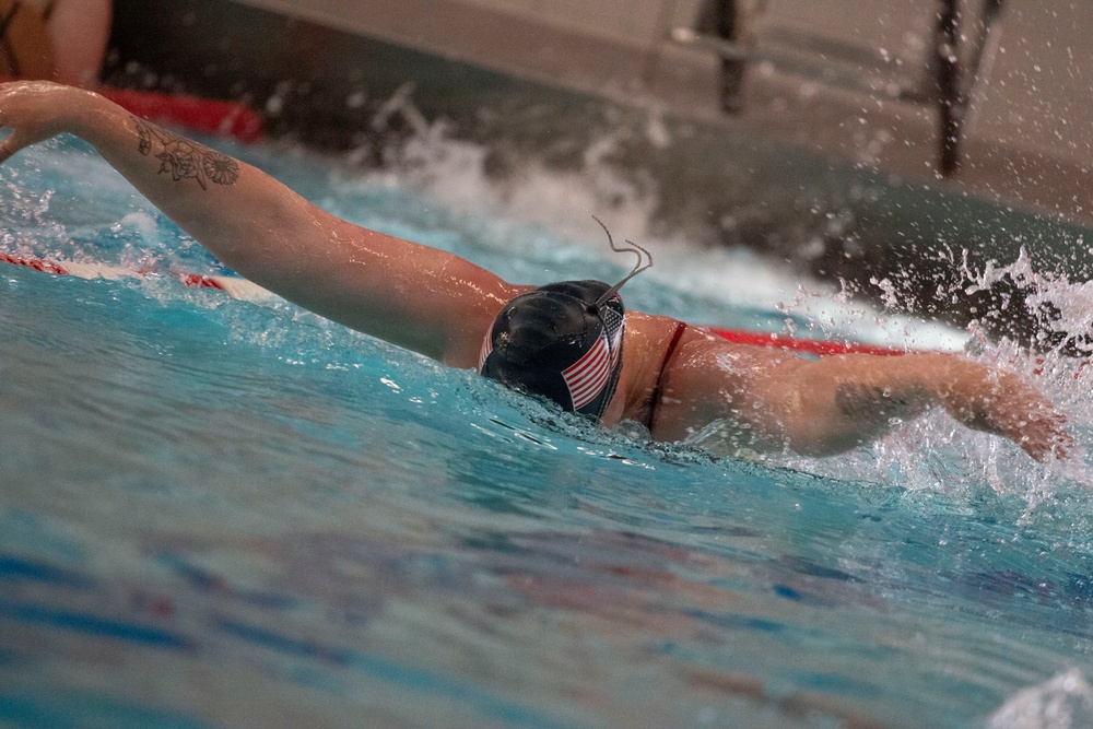 A competitive swimmer is performing the butterfly stroke in an indoor swimming pool. The swimmer is wearing a dark cap decorated with an American flag design and has visible tattoos on both arms. The image captures the dynamic motion of the stroke with significant water splash around the swimmer.