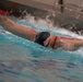 A competitive swimmer is performing the butterfly stroke in an indoor swimming pool. The swimmer is wearing a dark cap decorated with an American flag design and has visible tattoos on both arms. The image captures the dynamic motion of the stroke with significant water splash around the swimmer.