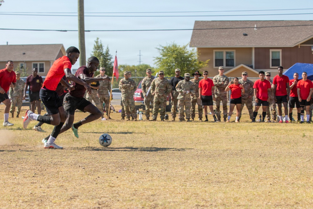 A soccer match is being played on a dry dirt field at what appears to be a military installation, with two players actively competing for the ball in the foreground. A large crowd of spectators, consisting of both uniformed military personnel in camouflage and players in red team uniforms, lines the sideline watching the action. Residential buildings and trees are visible in the background, consistent with on-base housing.