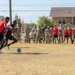 A soccer match is being played on a dry dirt field at what appears to be a military installation, with two players actively competing for the ball in the foreground. A large crowd of spectators, consisting of both uniformed military personnel in camouflage and players in red team uniforms, lines the sideline watching the action. Residential buildings and trees are visible in the background, consistent with on-base housing.