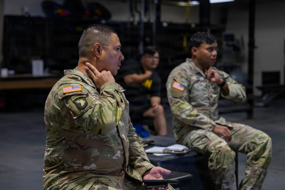 Two U.S. Army soldiers in OCP (Operational Camouflage Pattern) uniforms are seated and attentively listening, likely during a briefing or training session inside what appears to be a military fitness facility or gymnasium. The soldier in the foreground displays a 101st Airborne Division patch and an Airborne tab on his left shoulder. A third individual in civilian athletic wear is visible seated in the background.