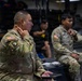 Two U.S. Army soldiers in OCP (Operational Camouflage Pattern) uniforms are seated and attentively listening, likely during a briefing or training session inside what appears to be a military fitness facility or gymnasium. The soldier in the foreground displays a 101st Airborne Division patch and an Airborne tab on his left shoulder. A third individual in civilian athletic wear is visible seated in the background.