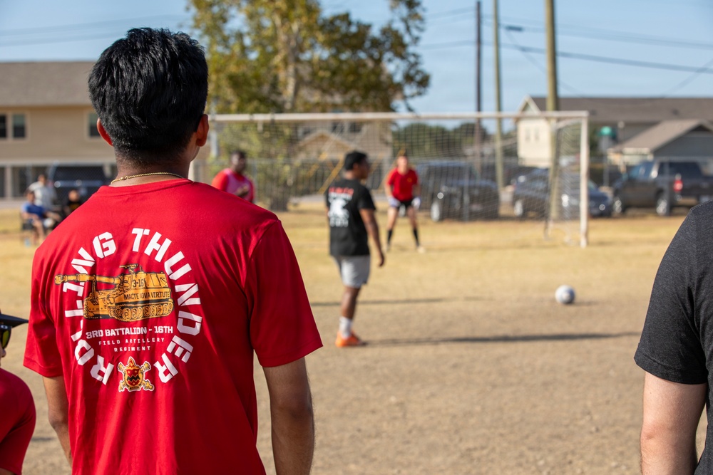A group of people are playing an informal soccer game on a dry grass field near a residential area. The foreground features a person wearing a red 'Rolling Thunder' military unit shirt from the 3rd Battalion, 16th Field Artillery Regiment. A soccer goal with netting is visible in the background along with parked vehicles and houses.