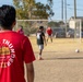 A group of people are playing an informal soccer game on a dry grass field near a residential area. The foreground features a person wearing a red 'Rolling Thunder' military unit shirt from the 3rd Battalion, 16th Field Artillery Regiment. A soccer goal with netting is visible in the background along with parked vehicles and houses.