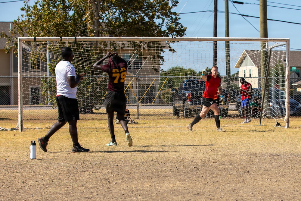 A casual outdoor soccer match is being played on a dry, grass-sparse field. A goalkeeper in a red sleeveless shirt is positioned in front of a white metal goal with netting, while players in dark jerseys and white shirts are nearby. The setting appears to be a neighborhood recreational area with buildings, trees, and utility poles visible in the background.