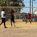 A casual outdoor soccer match is being played on a dry, grass-sparse field. A goalkeeper in a red sleeveless shirt is positioned in front of a white metal goal with netting, while players in dark jerseys and white shirts are nearby. The setting appears to be a neighborhood recreational area with buildings, trees, and utility poles visible in the background.