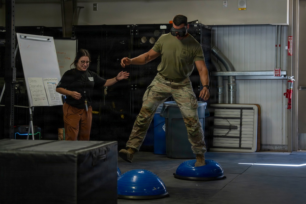 A person in U.S. Army military uniform and VR headset is balancing on a BOSU ball as part of a 'Sleep Deprivation Simulation' exercise. A female instructor in a black shirt and brown pants stands nearby with hands extended, ready to assist. The activity appears to be a controlled training demonstration taking place inside a gymnasium or training facility.