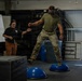 A person in U.S. Army military uniform and VR headset is balancing on a BOSU ball as part of a 'Sleep Deprivation Simulation' exercise. A female instructor in a black shirt and brown pants stands nearby with hands extended, ready to assist. The activity appears to be a controlled training demonstration taking place inside a gymnasium or training facility.