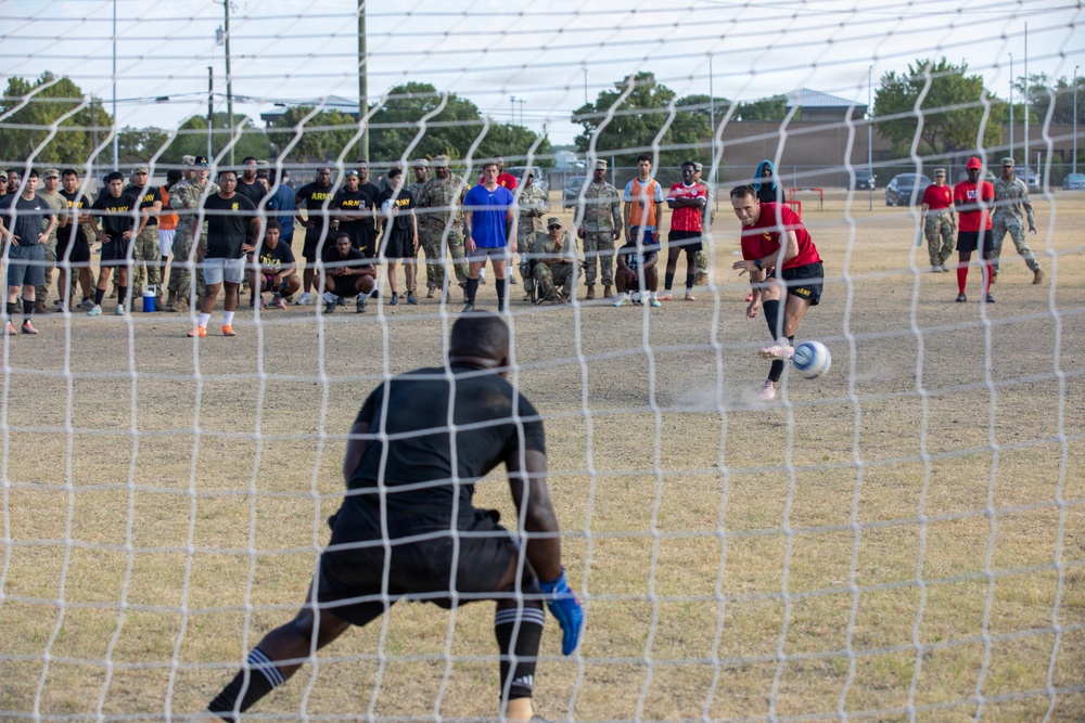 A soccer penalty kick is being taken on a dry grass field, viewed from behind the goal net. A player in a red jersey is kicking the ball toward a goalkeeper dressed in black. A large crowd of spectators, many wearing U.S. Army uniforms and PT gear, watches from the sideline.