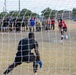 A soccer penalty kick is being taken on a dry grass field, viewed from behind the goal net. A player in a red jersey is kicking the ball toward a goalkeeper dressed in black. A large crowd of spectators, many wearing U.S. Army uniforms and PT gear, watches from the sideline.