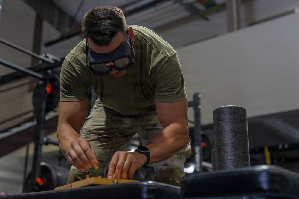 A person wearing military-style OCP (Operational Camouflage Pattern) pants and an olive drab t-shirt is leaning over a surface, working with small objects on a wooden board. They have protective goggles pushed up on their forehead and are wearing a smartwatch. The setting appears to be an indoor facility, possibly a military gym or workshop.