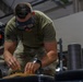A person wearing military-style OCP (Operational Camouflage Pattern) pants and an olive drab t-shirt is leaning over a surface, working with small objects on a wooden board. They have protective goggles pushed up on their forehead and are wearing a smartwatch. The setting appears to be an indoor facility, possibly a military gym or workshop.