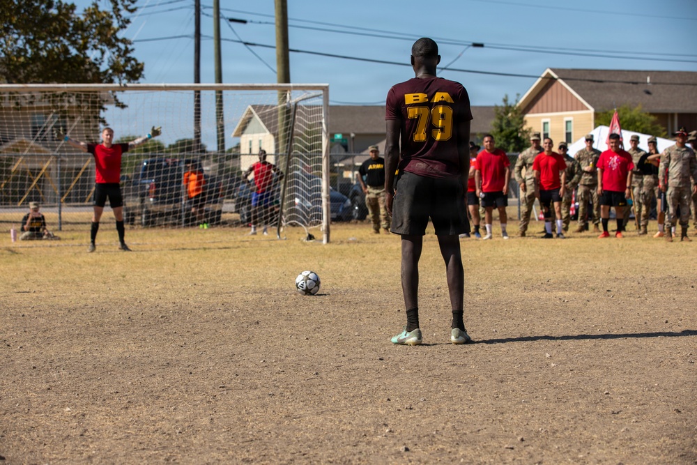 A soccer player wearing jersey number 79 with 'BA' lettering stands facing a goal, preparing to take a penalty kick on a dirt field. A goalkeeper is positioned in the goal with arms outstretched, while a crowd of spectators — many appearing to be in military uniforms — lines the sideline. The setting appears to be a recreational or intramural soccer match at a military installation.